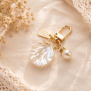 May include: A gold-colored keychain with a pearlescent, shell-shaped charm and a dangling faux pearl. The keychain also features a small rectangular tag with text. The background is a soft, neutral color.