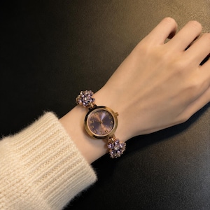 May include: A gold-tone wristwatch featuring a round face with a purple dial and gold numerals. The watch band is adorned with multiple purple flower-shaped accents, each set with small clear stones, creating a decorative link bracelet design.