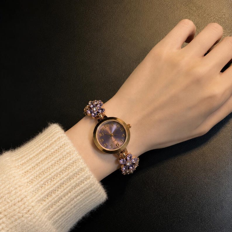 May include: A gold-tone wristwatch featuring a round face with a purple dial and gold numerals. The watch band is adorned with multiple purple flower-shaped accents, each set with small clear stones, creating a decorative link bracelet design.