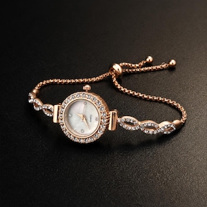 May include: A rose gold-tone quartz watch with a round mother-of-pearl dial, featuring a bezel encrusted with clear crystals. The bracelet consists of twisted metal links set with crystals and an adjustable sliding clasp mechanism.