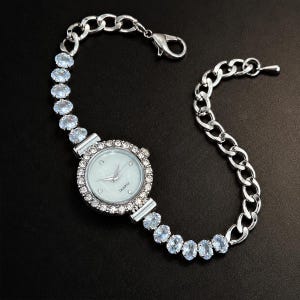 Puede incluir: Un reloj de cuarzo plateado con esfera redonda blanca, manecillas plateadas y bisel con cristales transparentes. La pulsera combina piedras ovaladas de color azul claro con eslabones de cadena plateados y un cierre de mosquetón.