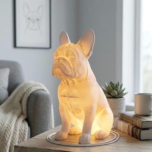 May include: A white ceramic bulldog lamp sits on a wooden table. The lamp is lit from within, casting a warm glow. A framed sketch of a bulldog hangs on the wall behind the lamp. The lamp is a decorative home decor item.