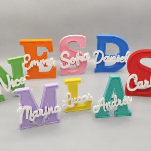 Op de afbeelding: Een verzameling kleurrijke, 3D-geprinte naamborden. Elk bord heeft een grote, vette letter in een andere kleur, met een kleinere, cursieve naam in wit. De namen zijn Nico, Emma, Sofia, Daniel, Carla, Marina, Lucas en Andrea.