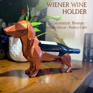 May include: A geometric bronze wine bottle holder in the shape of a dachshund dog. The holder is designed to cradle a wine bottle. The text on the image reads "THE LOW-POLY WIENER WINE HOLDER".