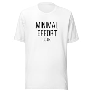 Puede incluir: Camiseta blanca con el texto negro "MINIMAL EFFORT CLUB" centrado en la parte delantera. El texto está en negrita y sin serifa. La camiseta tiene cuello redondo y mangas cortas.