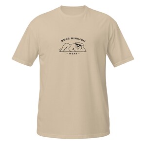 May include: A tan t-shirt featuring a black graphic of a bear wearing sunglasses, with the words "BEAR MINIMUM" above the bear and "-WEAR-" below. The shirt has short sleeves and a crew neck.