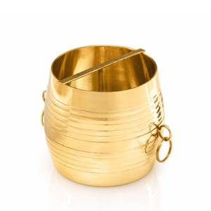 May include: A gold-colored metal ice bucket with a horizontal bar across the top. The bucket has a cylindrical shape with horizontal ridges and two ring handles on the side. The ice bucket is on a white background.