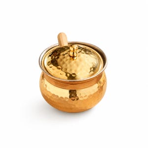 May include: A small, gold-colored hammered metal sugar bowl with a lid and a wooden handle. The bowl has a round shape and a silver rim. The lid is also gold and has a textured surface.