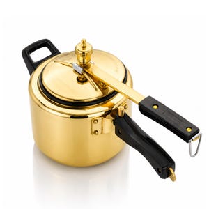 Puede incluir: Una olla a presión de color dorado con un acabado metálico pulido. Cuenta con una tapa de cierre seguro con una válvula de presión central, un mango largo negro unido a la tapa y un mango lateral negro más corto en el cuerpo cilíndrico.