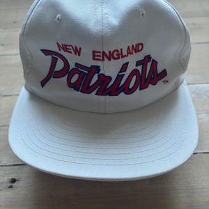 Może przedstawiać: Biała czapka bejsbolowa z napisem "NEW ENGLAND Patriots" w czerwonym i niebieskim skrypcie. Czapka ma strukturalną koronę i zakrzywiony daszek, z widocznymi detalami szwów. Klasyczny dodatek sportowy.