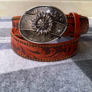 May include: A brown leather belt with a sunflower buckle. The belt features embossed floral patterns and brass-colored rivets along the edges. The buckle is silver-toned with a detailed sunflower design.