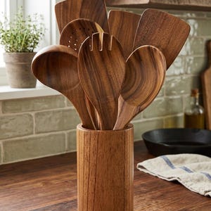 Könnte beinhalten: Ein Set aus hölzernen Küchenutensilien, bestehend aus einem Schaumlöffel, einem Löffel, einer Gabel und flachen Pfannenwendern, die in einem zylindrischen Utensilienhalter aus Holz stehen. Die Teile haben eine natürliche braune Holzmaserung.