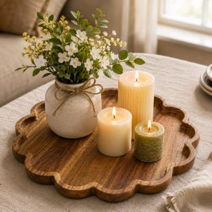 May include: A round wooden tray with a scalloped edge holds a cream-coloured ceramic vase containing white and yellow artificial flowers tied with twine, alongside three lit pillar candles in varying heights and shades of cream and sage green.