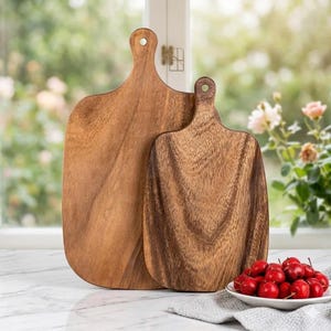 Peut inclure: Deux planches à découper en bois brun avec des motifs de grain visibles et des poignées arrondies dotées de petits trous circulaires au sommet. Les planches sont placées verticalement, l'une derrière l'autre, avec une surface lisse et polie.