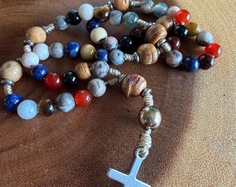 May include: A rosary featuring a variety of multicolored beads including wood, stone, and glass in shades of blue, red, white, black, and brown. The beads are strung on light-colored cord with a single metallic bead and a silver-toned metal cross.