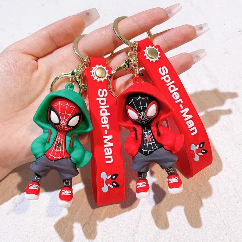 Może przedstawiać: Dwa breloczki do kluczy Spider-Man. Jeden breloczek przedstawia zieloną postać Spider-Mana z kapturem, a drugi czerwoną postać Spider-Mana z kapturem. Oba mają czerwone paski z napisem "Spider-Man".
