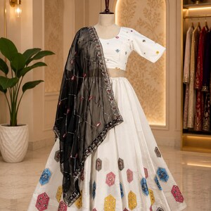 May include: A white lehenga choli set featuring a cropped blouse with short sleeves and a full-length skirt, both adorned with colourful hexagonal embroidered patches. A sheer black dupatta with silver grid embroidery and sequins is draped over.