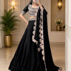 May include: A black lehenga choli set featuring a cropped blouse with intricate multi-colored embroidery and short sleeves. The matching floor-length skirt is solid black with a wide hem. A sheer black dupatta with a scalloped floral border completes the set.