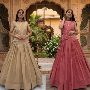 May include: Two sleeveless outfits consisting of a top and a long, pleated skirt. One outfit is beige with subtle embroidery, and the other is a dusty rose color with white floral embroidery on the top. Both feature a wide hem on the skirt.