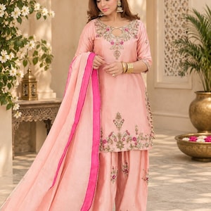 May include: A peach-colored salwar kameez set featuring a tunic with a keyhole neckline, three-quarter sleeves, and intricate floral embroidery in pink and gold thread. The matching wide-leg trousers and a sheer dupatta with a bright pink border.