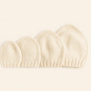 Może przedstawiać: Zestaw czterech kremowych dzianinowych czapek typu beanie ułożonych w rzędzie od najmniejszej do największej. Każda czapka ma prosty wzór ściegu pończoszniczego na korpusie oraz ściągacz na dole, wykonane z miękkiej, neutralnej przędzy.