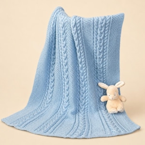 May include: A light blue knitted blanket featuring vertical cable knit patterns and textured seed stitch panels. A small, cream-colored plush rabbit toy with long ears is positioned on the lower right side of the folded fabric.