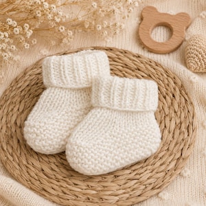 May include: A pair of cream-coloured knitted baby booties featuring a garter stitch foot and a ribbed cuff. The booties are crafted from a soft yarn and display a simple, clean design with a folded top edge, showcasing a classic texture.