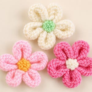 以下が含まれることがあります： 5枚の花びらを持つかぎ針編みの花が3つ。上の花はクリーム色で中心がライトグリーン。左下の花はライトピンクで中心が黄色。右下の花はダークピンクで中心がクリーム色です。