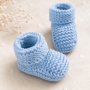 May include: Two light blue knitted baby booties. The booties are made of soft yarn and have a rolled-down cuff. The booties are displayed on a cream-coloured, textured surface. The booties are a charming and cosy accessory for newborns.