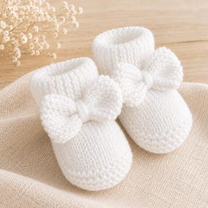 May include: A pair of white knitted baby booties featuring a textured stitch pattern and a prominent decorative bow on the front of each shoe. The booties have a soft, rounded toe design and a ribbed cuff at the top, crafted from a solid white yarn.
