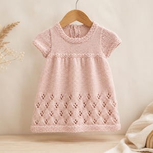 Pode incluir: Um vestido de bebê de malha rosa claro com mangas curtas, pendurado em um cabide de madeira. O vestido apresenta um corpete texturizado e uma saia com padrão de diamante. O vestido é feito de fio macio e é adequado para um bebê.