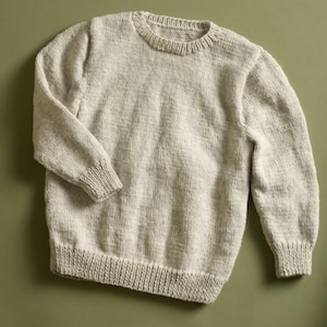 May include: A long-sleeved crew neck sweater in a light oatmeal beige color. The garment features a ribbed knit pattern at the collar, cuffs, and hem, with a consistent stockinette stitch texture throughout the body and sleeves of the knit pullover.