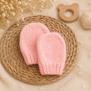 Puede incluir: Un par de manoplas de punto para bebé en color rosa claro, con una forma redondeada sencilla y un puño acanalado. Las manoplas están confeccionadas con hilo texturizado y se presentan juntas, mostrando un patrón de tejido uniforme en toda la prenda.