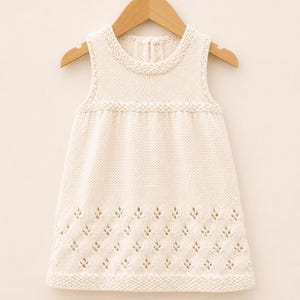 Pode incluir: Um vestido de malha sem mangas na cor creme, com decote redondo e borda em ponto musgo. O corpo é liso, enquanto a parte inferior apresenta um delicado padrão rendado repetitivo em forma de folhas, finalizado com uma bainha em ponto musgo.