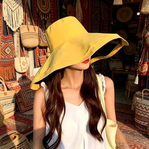 Puede incluir: Un sombrero de sol amarillo de ala ancha con un interior oscuro. La persona lleva el sombrero con pelo largo y casta&ntilde;o y una camiseta blanca. El fondo presenta varias bolsas y textiles, lo que sugiere una tienda o mercado.