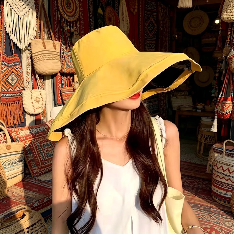 Puede incluir: Un sombrero de sol amarillo de ala ancha con un interior oscuro. La persona lleva el sombrero con pelo largo y casta&ntilde;o y una camiseta blanca. El fondo presenta varias bolsas y textiles, lo que sugiere una tienda o mercado.