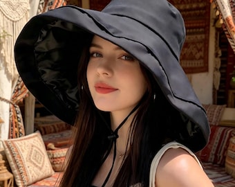 Puede incluir: Un sombrero negro para el sol con ala ancha y cordón. El sombrero tiene un diseño estructurado y parece estar hecho de una tela lisa. El sombrero lo lleva una persona con pelo largo y oscuro.