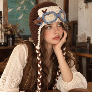 May include: A brown and cream crocheted aviator hat featuring a pair of light blue crocheted goggles attached to the front. The hat includes long, braided earflaps made of alternating brown and cream yarn, extending down from the sides.