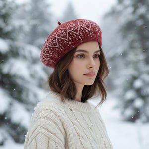 May include: A deep red knit beret featuring a white geometric fair isle pattern and a small top pom-pom. The hat is worn with a cream-colored cable knit sweater, showcasing a textured wool construction and a classic rounded silhouette.
