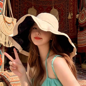 Puede incluir: Un sombrero para el sol de ala ancha con exterior color crema y parte inferior negra. El sombrero lo lleva una persona. La persona lleva una camiseta verde claro. El fondo incluye un bolso tejido y un tapiz estampado.