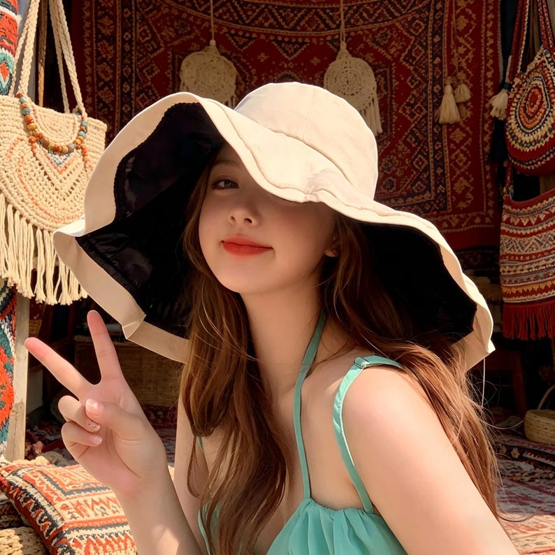 Puede incluir: Un sombrero para el sol de ala ancha con exterior color crema y parte inferior negra. El sombrero lo lleva una persona. La persona lleva una camiseta verde claro. El fondo incluye un bolso tejido y un tapiz estampado.