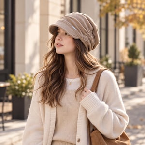 May include: A beige corduroy newsboy cap featuring a gathered, slouchy crown and a short, stiff brim. The hat includes a small, rectangular silver-toned metal accent on the side, providing a textured, soft appearance with a structured front visor.
