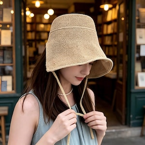 May include: A beige woven sun hat with a wide brim and a structured crown. The hat features a chin strap and is designed to provide shade. The hat is made of a natural fiber material, perfect for outdoor wear.