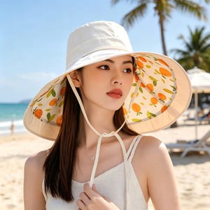 May include: A white sun hat with a wide brim, featuring an orange fruit pattern on the underside. The hat has a chin strap and is worn by a person on a sunny beach. The hat is designed to provide sun protection.