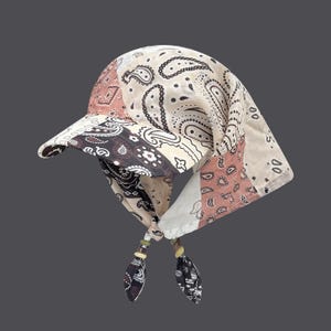 May include: A bandana-style hat with a visor, featuring a patchwork design in shades of beige, brown, and pink. The fabric is patterned with paisley motifs. The hat has ties with decorative beads.