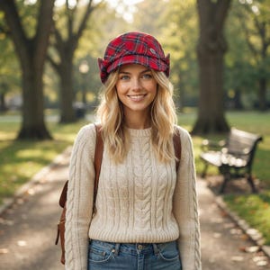 May include: A red and blue plaid cap with ear flaps and a small silver button on the side, worn with a beige cable-knit sweater and blue denim jeans. A brown leather backpack is visible over the shoulders of the sweater.