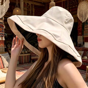 Puede incluir: Un sombrero para el sol de color crema con ala ancha, interior oscuro y un sutil ribete gris. El sombrero es sostenido, mostrando su forma y dise&ntilde;o. El sombrero parece estar hecho de un material ligero, ideal para la protecci&oacute;n solar.