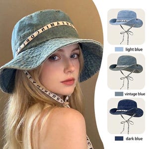May include: A denim bucket hat with a wide brim and a white ribbon band featuring the text DREAM. The hat includes long, dangling ribbon ties with the same text. Three additional color variations are shown: light blue, vintage blue, and dark blue.
