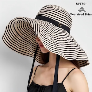 May include: A wide-brimmed sun hat with black and cream horizontal stripes. The hat features a black band and ties, and the text "UPF50+" and "Oversized Brim" are visible. The hat is designed for sun protection.