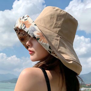 以下が含まれることがあります： ベージュの日よけ帽子、つば広。 つばの裏側には、オレンジとクリーム色の花柄が施されています。 この帽子は日焼け防止用に設計されており、アウトドアアクティビティに適しています。